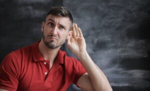 A man in red polo short standing in front of a chalkboard with his hand raised to his ear as if listening carefully with a skeptical look on his face. Negative Feedback?