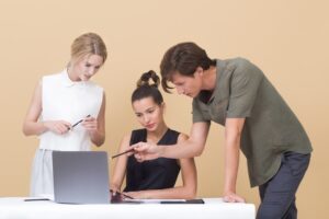 Three people gathered around a computer with one seated. Two are providing negative feedback and positive feedback.